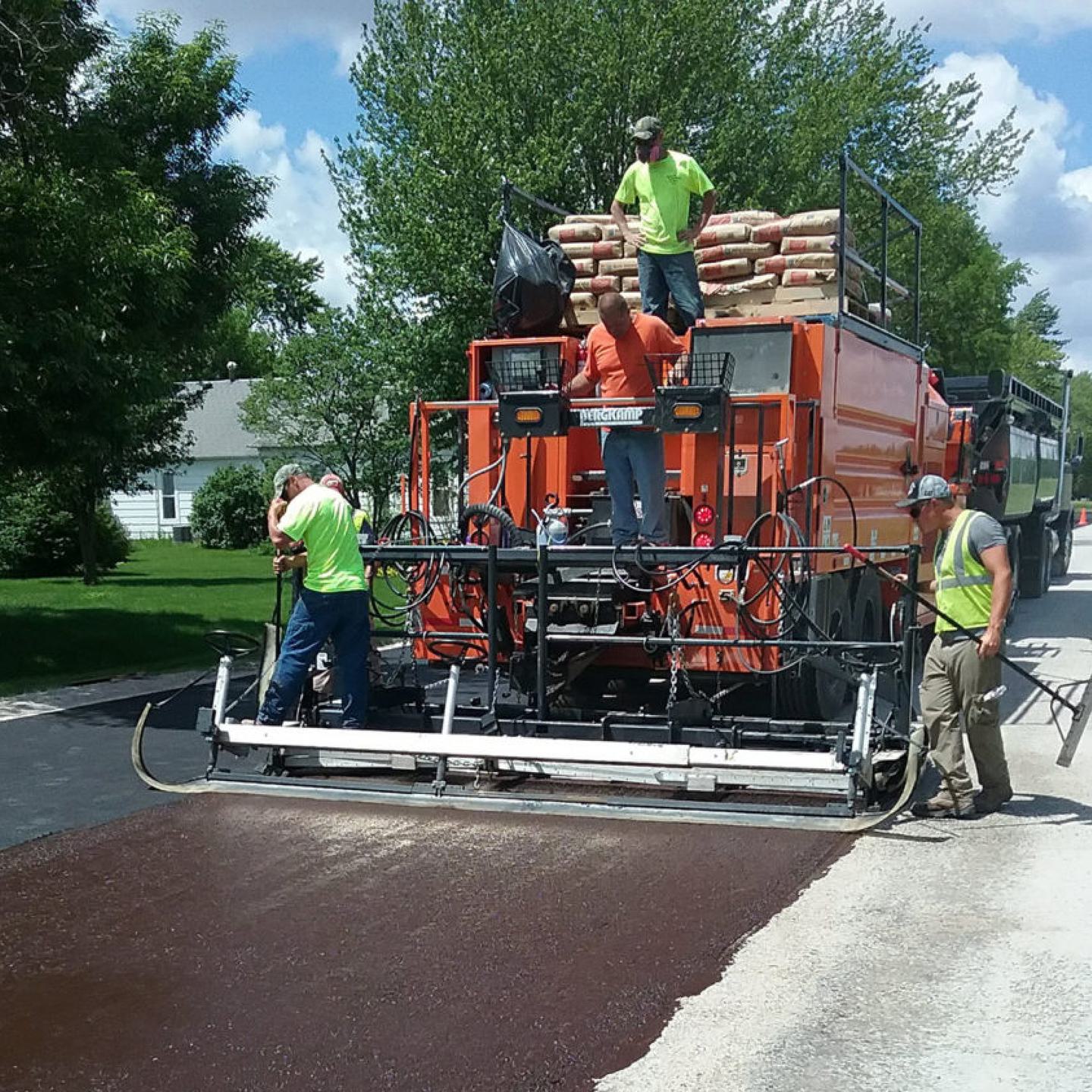 Orange truck-mounted slurry seal and microsurfacing paver applying a dark mat on a residential street with handwork at the edge