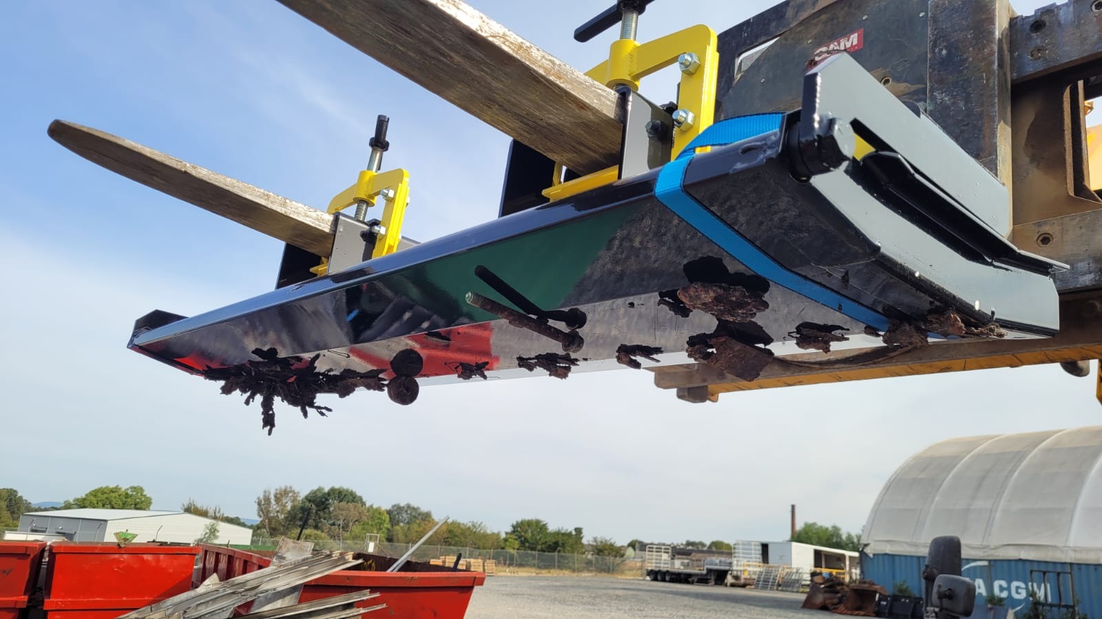 Black forklift-mounted magnetic sweeper raised above a yard with rusty bolts and scrap clinging to the magnet face