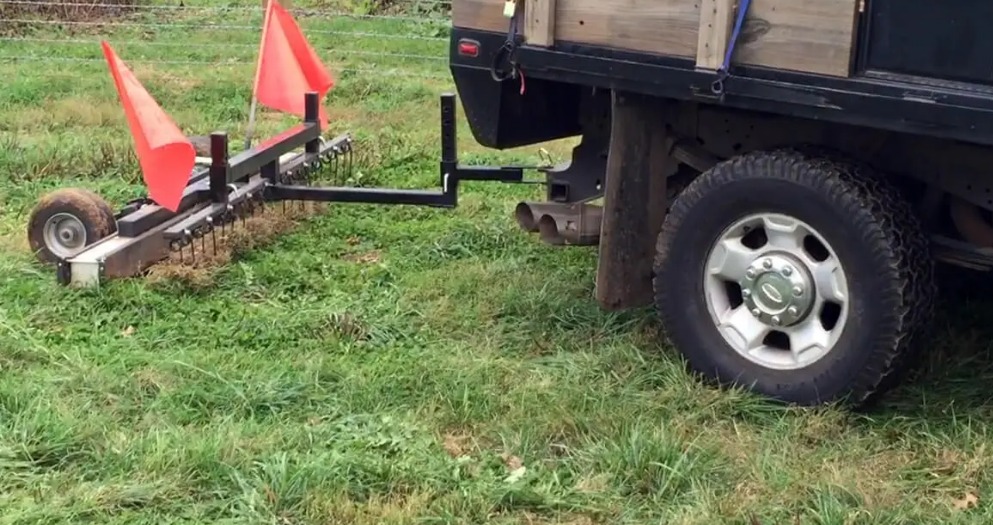 Black tow-behind magnetic sweeper with orange safety flags hitched to a pickup truck on grass