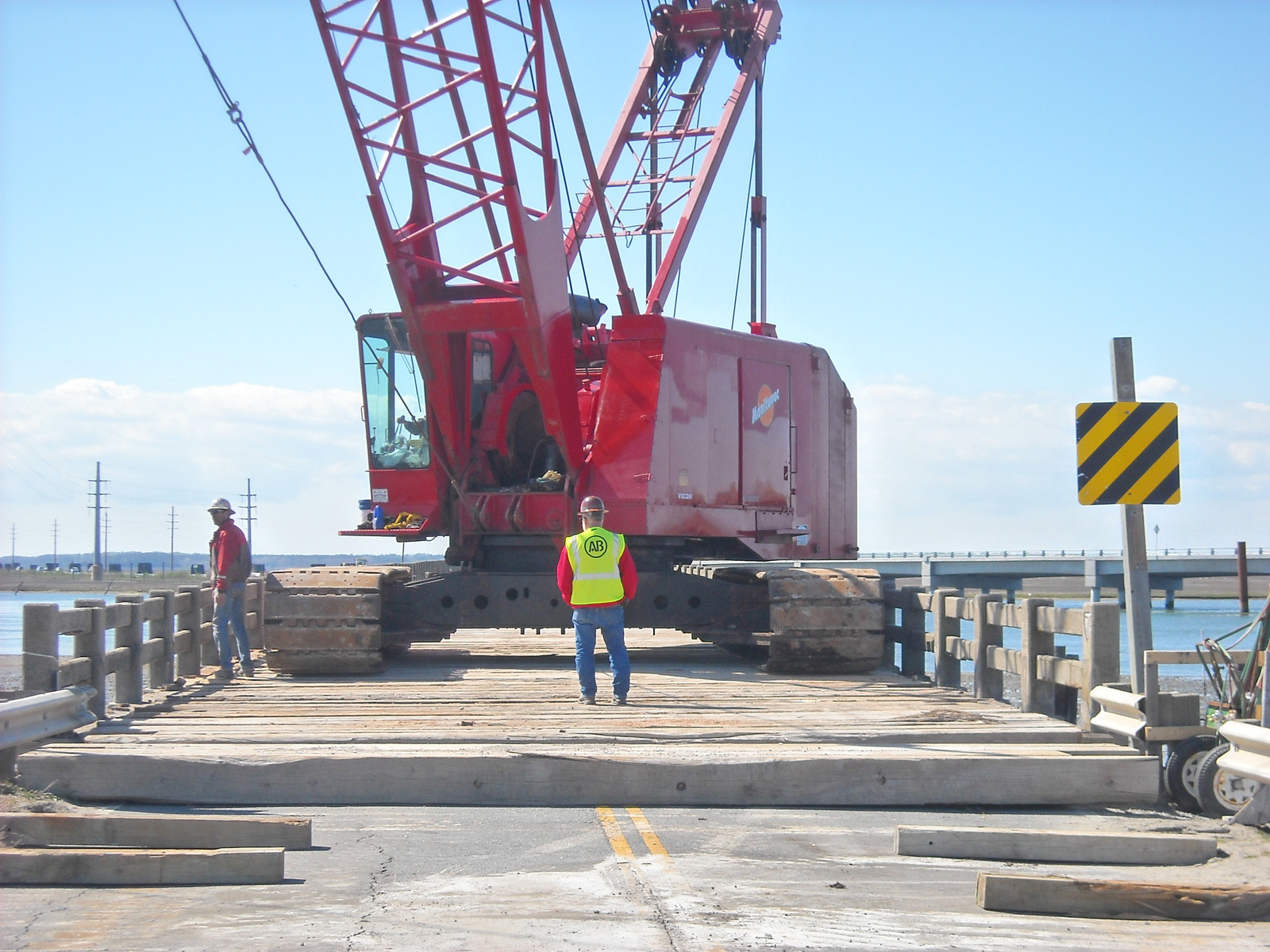 Red Manitowoc crawler crane with tracks on timber crane mats laid across a bridge roadway with workers spotting the walk
