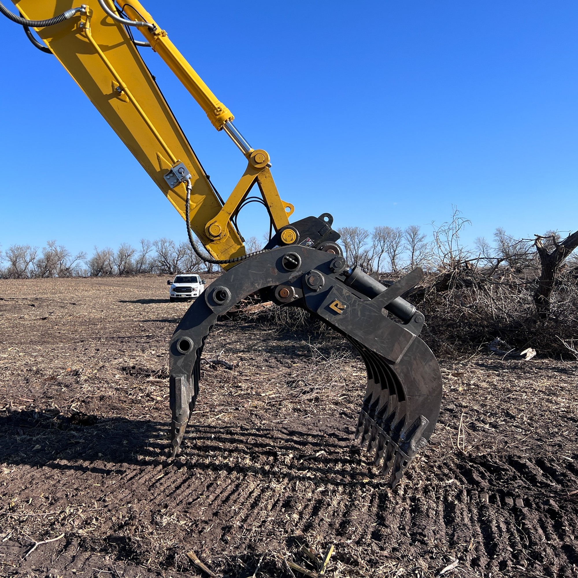 Dark Rockland rake grapple on yellow excavator arm at a land-clearing site with brush pile