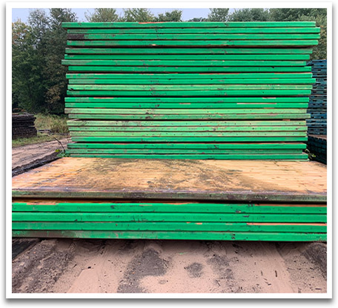 Tall outdoor stack of laminated wood access mats with green painted edges on sandy ground