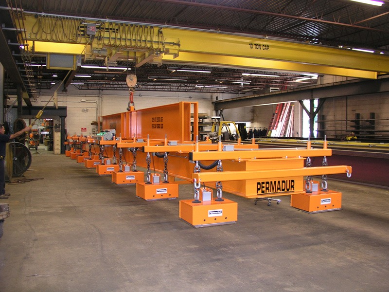 Large orange multi-magnet Permadur plate lifting beam suspended from a yellow overhead crane in a fabrication shop