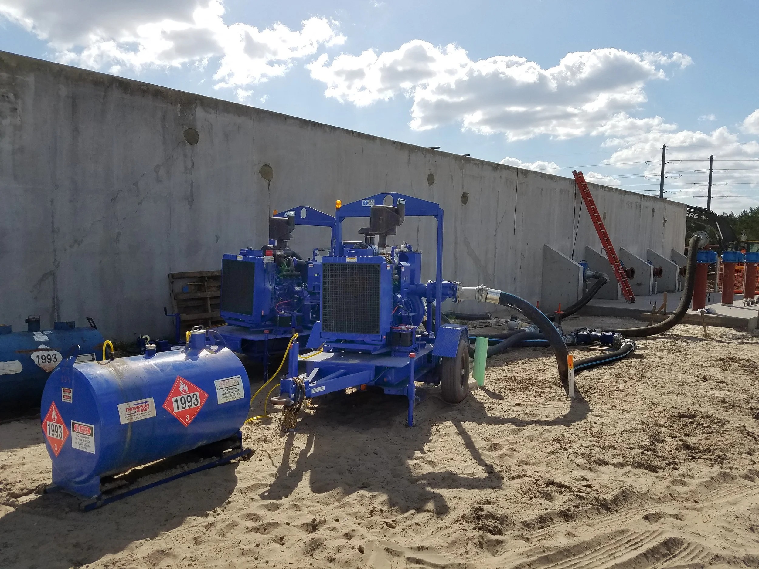 Two blue trailer-mounted pumps and diesel fuel tanks with hoses connected on a sandy construction site