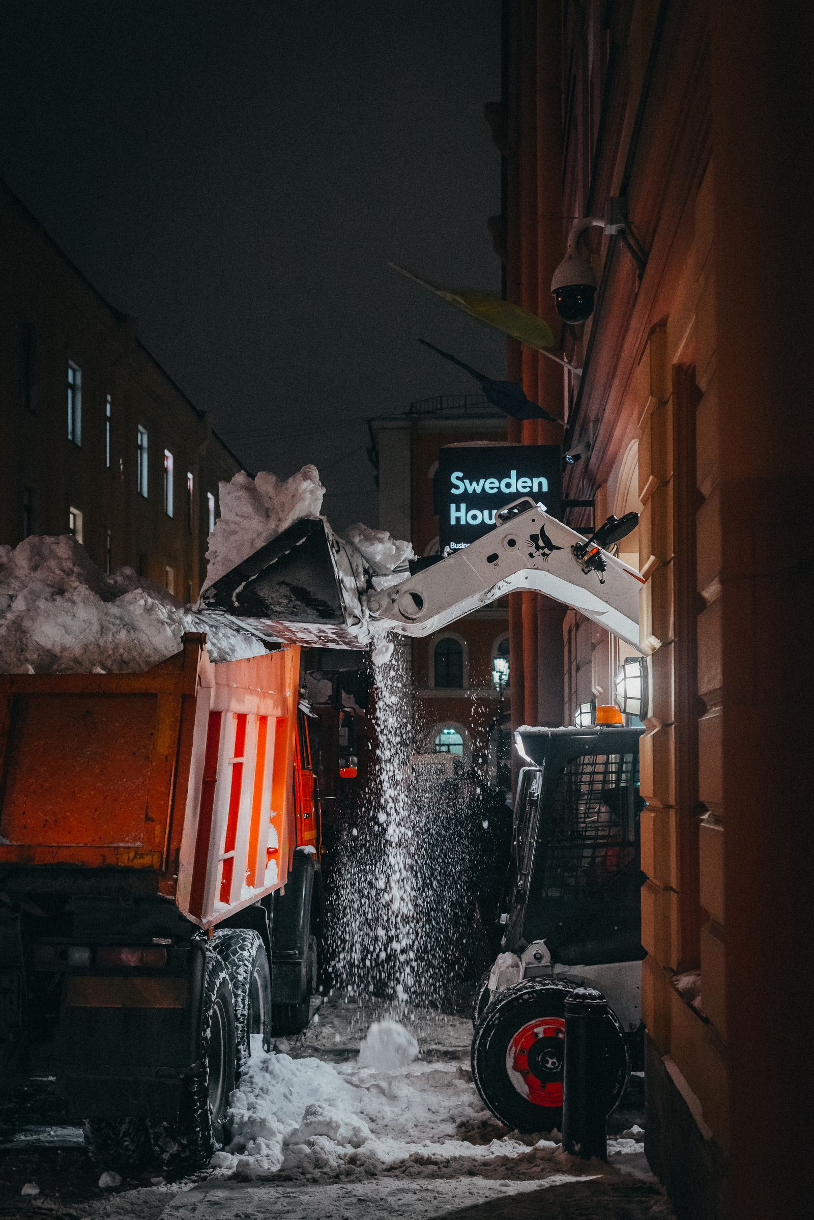 Skid steer dumping light snow into a truck bed during night cleanup