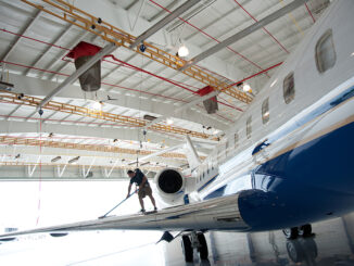 Technician in a harness on a jet wing connected to yellow overhead rigid-rail fall protection tracks in an aircraft hangar