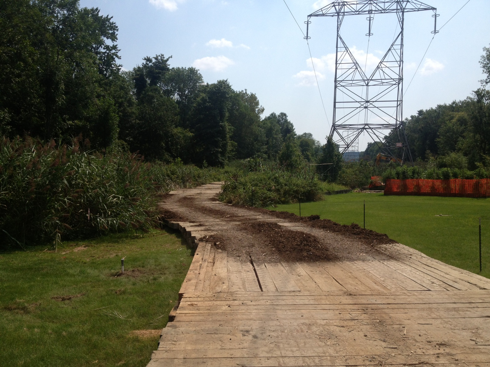 Temporary access road of layered timber crane mats with longitudinal bearing paths through marsh grass toward a high-voltage transmission tower