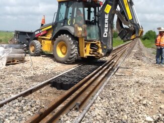 Yellow backhoe loader with tire on black textured ground mat over railroad ballast while a worker in hi-vis observes
