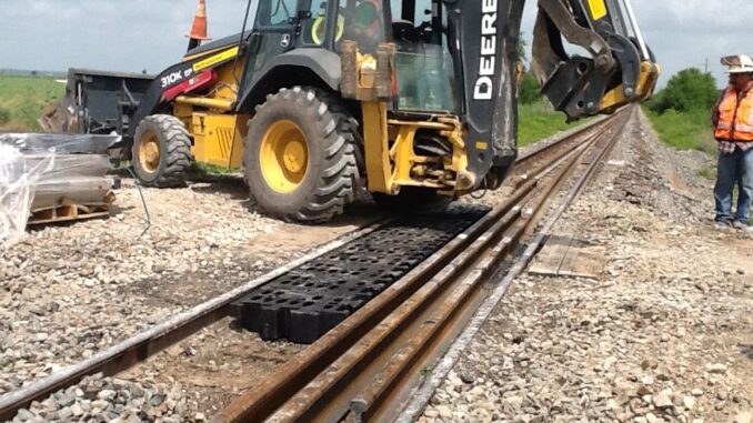 Yellow backhoe loader with tire on black textured ground mat over railroad ballast while a worker in hi-vis observes