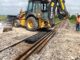 Yellow backhoe loader with tire on black textured ground mat over railroad ballast while a worker in hi-vis observes