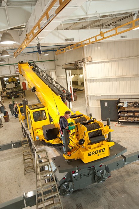 Worker in a harness on a Grove mobile crane connected to a yellow ceiling-mounted rigid-rail fall protection monorail in a shop