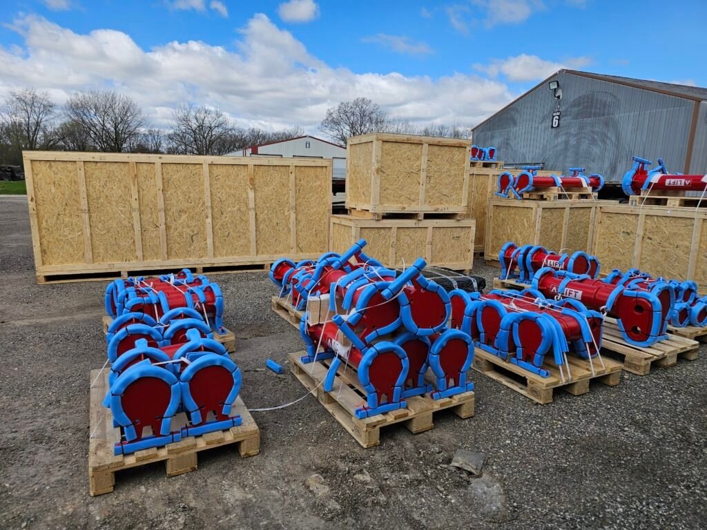 Red industrial lifting components with blue protective padding staged on wooden pallets in a gravel yard beside shipping crates