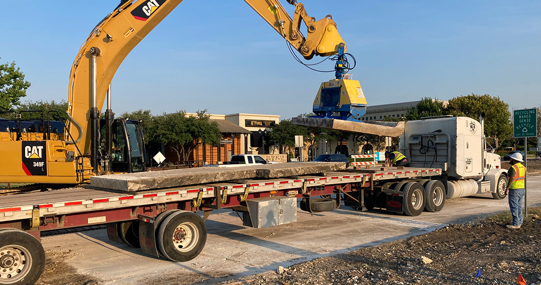 Excavator with vacuum attachment lifting a precast concrete slab from a flatbed trailer without slings
