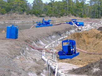 Blue skid pumps with white wellpoint header pipe and flexible hoses on a dirt embankment beside open water