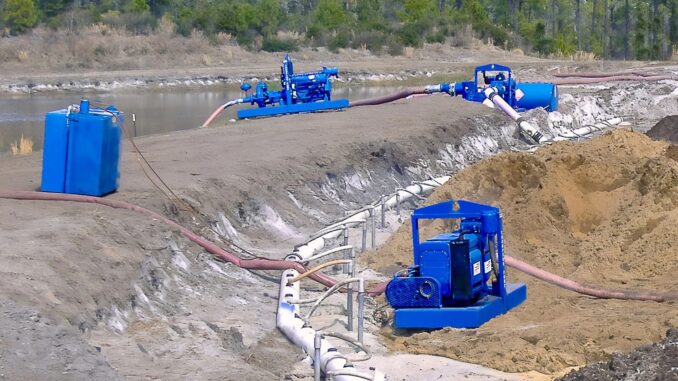 Blue skid pumps with white wellpoint header pipe and flexible hoses on a dirt embankment beside open water