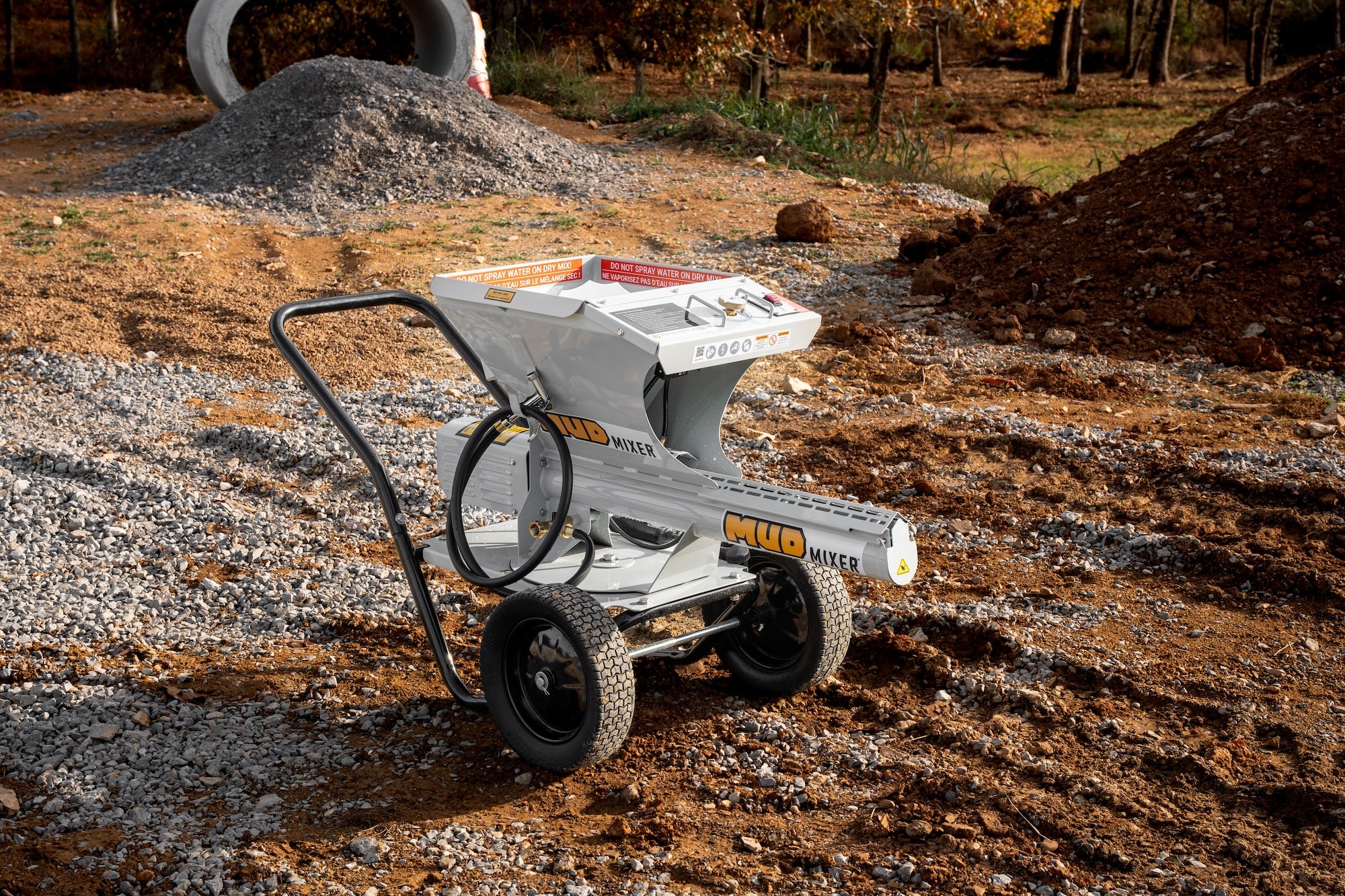 Light gray MudMixer portable continuous concrete mixer parked on dirt and gravel at an outdoor construction site with trees behind