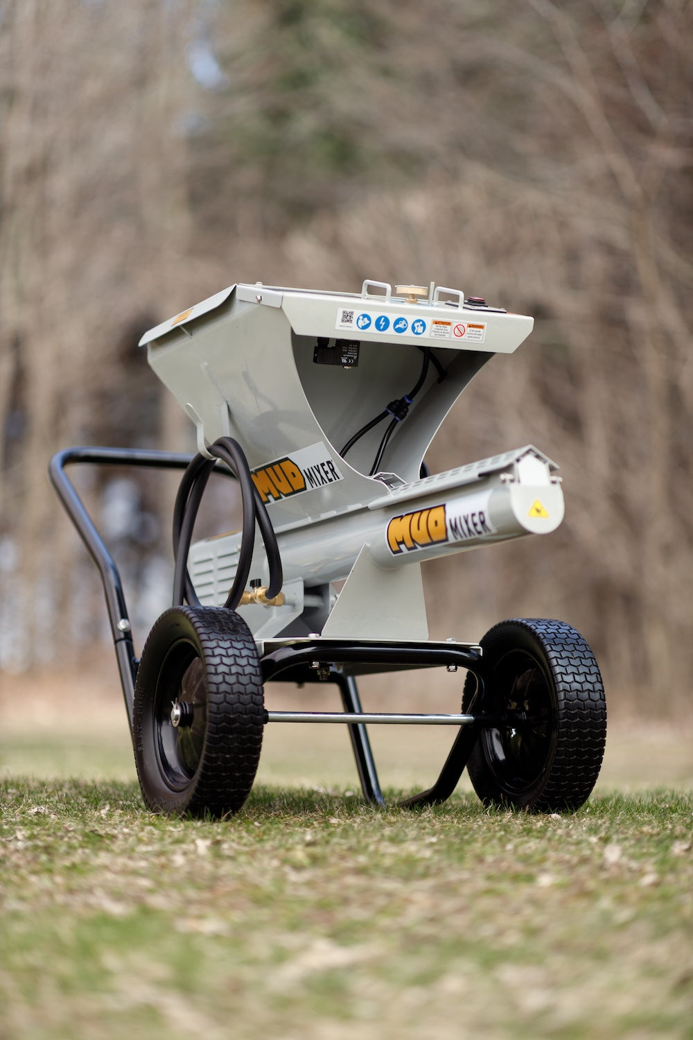 Three-quarter outdoor view of a gray MudMixer continuous concrete mixer with hopper, chute, and pneumatic tires on grass against blurred trees