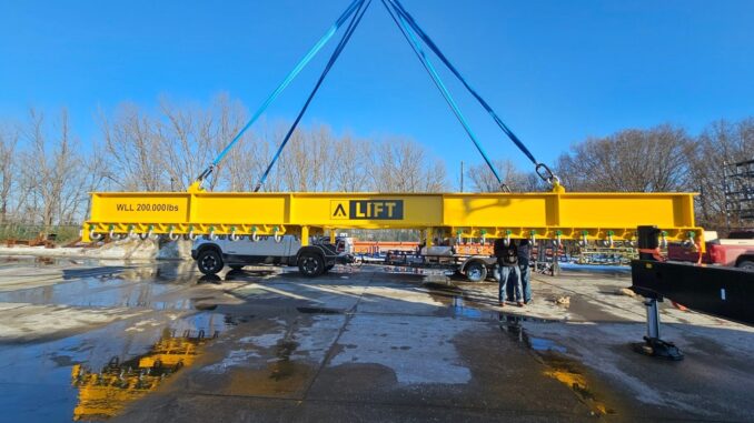 Yellow A-Lift spreader beam suspended on blue slings over wet concrete yard with workers