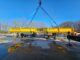 Yellow A-Lift spreader beam suspended on blue slings over wet concrete yard with workers