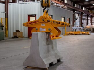 Yellow Tandemloc concrete barrier grab clamped on a gray Jersey barrier suspended from a crane hook in a warehouse