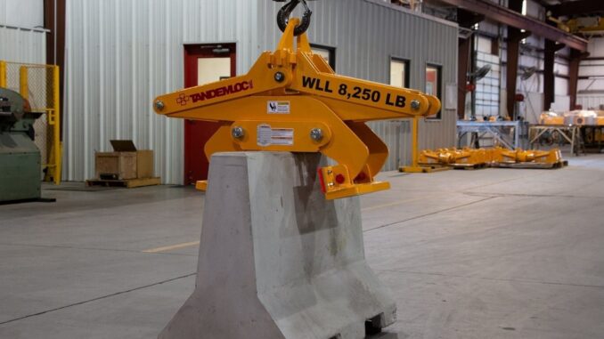 Yellow Tandemloc concrete barrier grab clamped on a gray Jersey barrier suspended from a crane hook in a warehouse