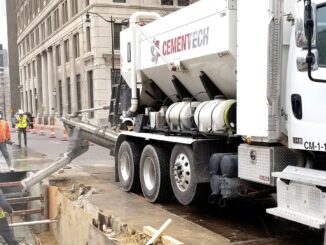 White Cemen Tech volumetric mixer truck pouring concrete into a city street excavation with crew nearby