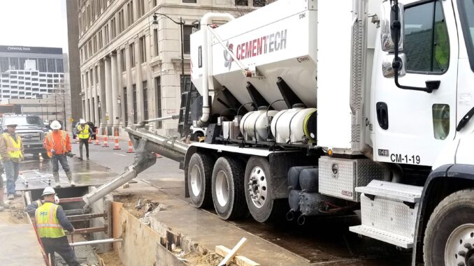 White Cemen Tech volumetric mixer truck pouring concrete into a city street excavation with crew nearby