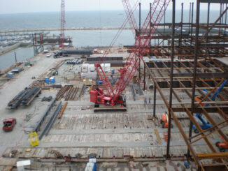 Red lattice-boom crawler crane working on a continuous timber crane-mat deck beside steel erection and waterfront staging