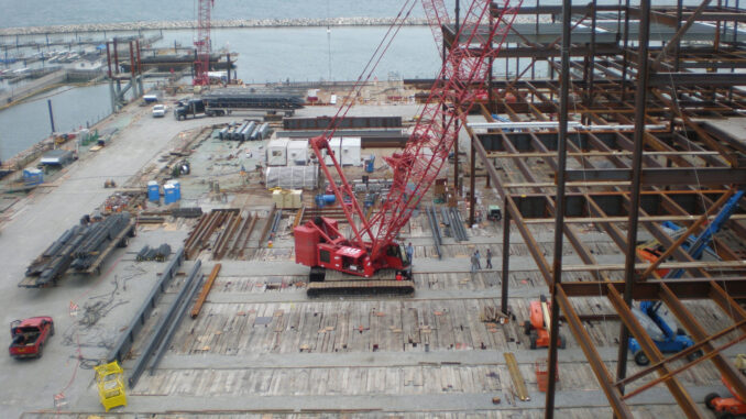 Red lattice-boom crawler crane working on a continuous timber crane-mat deck beside steel erection and waterfront staging