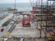 Red lattice-boom crawler crane working on a continuous timber crane-mat deck beside steel erection and waterfront staging