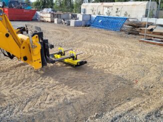 Yellow forklift-mounted magnetic sweeper hovering over gravel in an industrial yard with scrap bins and concrete blocks