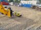 Yellow forklift-mounted magnetic sweeper hovering over gravel in an industrial yard with scrap bins and concrete blocks