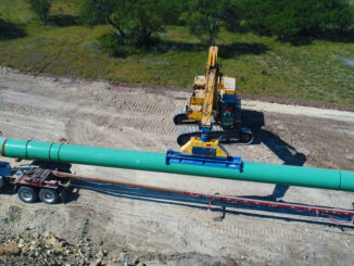 Excavator with vacuum lifter unloading large-diameter coated pipe from a flatbed trailer on a pipeline right-of-way
