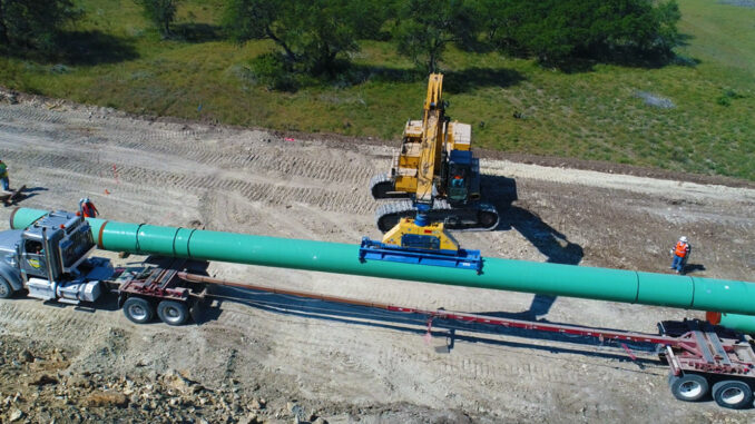 Excavator with vacuum lifter unloading large-diameter coated pipe from a flatbed trailer on a pipeline right-of-way