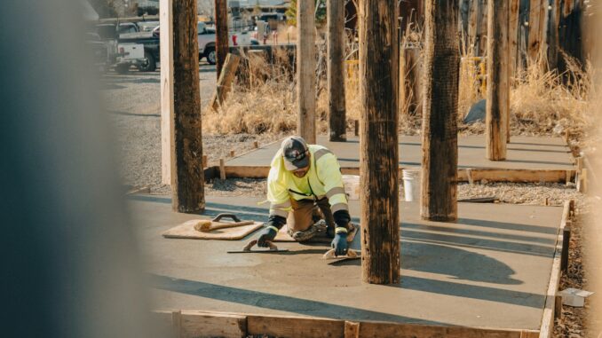 Worker finishing a freshly poured concrete slab between wood forms on a gravel lot with pickup trucks in the background