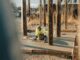 Worker finishing a freshly poured concrete slab between wood forms on a gravel lot with pickup trucks in the background