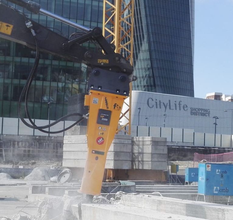 Yellow Indeco HP 2500 hydraulic breaker on an excavator arm breaking a concrete slab at an urban construction site