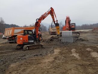 Orange Hitachi compact excavator with yellow Indeco hydraulic breaker working muddy ground beside dump trucks and a larger excavator