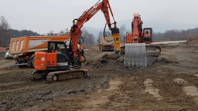 Orange Hitachi compact excavator with yellow Indeco hydraulic breaker working muddy ground beside dump trucks and a larger excavator