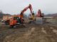 Orange Hitachi compact excavator with yellow Indeco hydraulic breaker working muddy ground beside dump trucks and a larger excavator